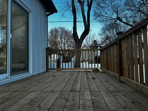 Wooden backyard deck with plank flooring, side railing, exterior wall, and trees visible beyond the deck.