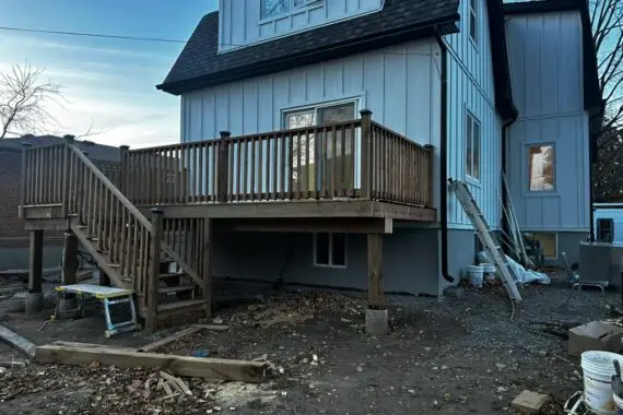 Residential house exterior with a second-floor wooden deck, stairs, railing, vertical siding, and construction materials visible below.