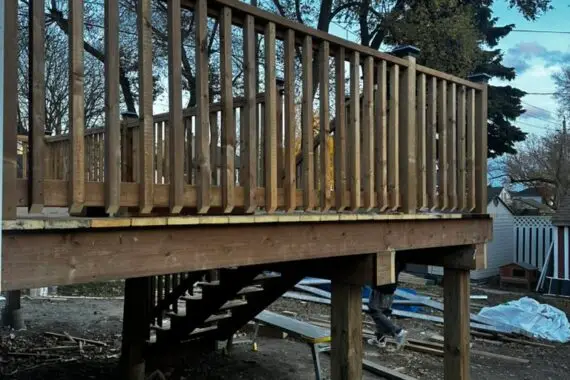 wooden deck under construction with railing, stairs, support posts, and unfinished ground below.