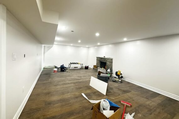 Basement renovation in progress with newly installed hardwood flooring, white painted walls, recessed ceiling lights, fireplace, and construction tools on site.