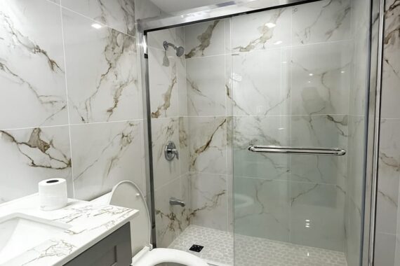 Modern bathroom with marble-look wall and floor tiles, glass shower enclosure, hexagon shower floor tiles, toilet, vanity, and recessed ceiling lighting.