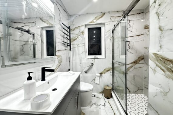 Modern bathroom renovation with marble-look wall tiles, glass shower enclosure, hexagon shower floor tiles, floating vanity, and black fixtures.