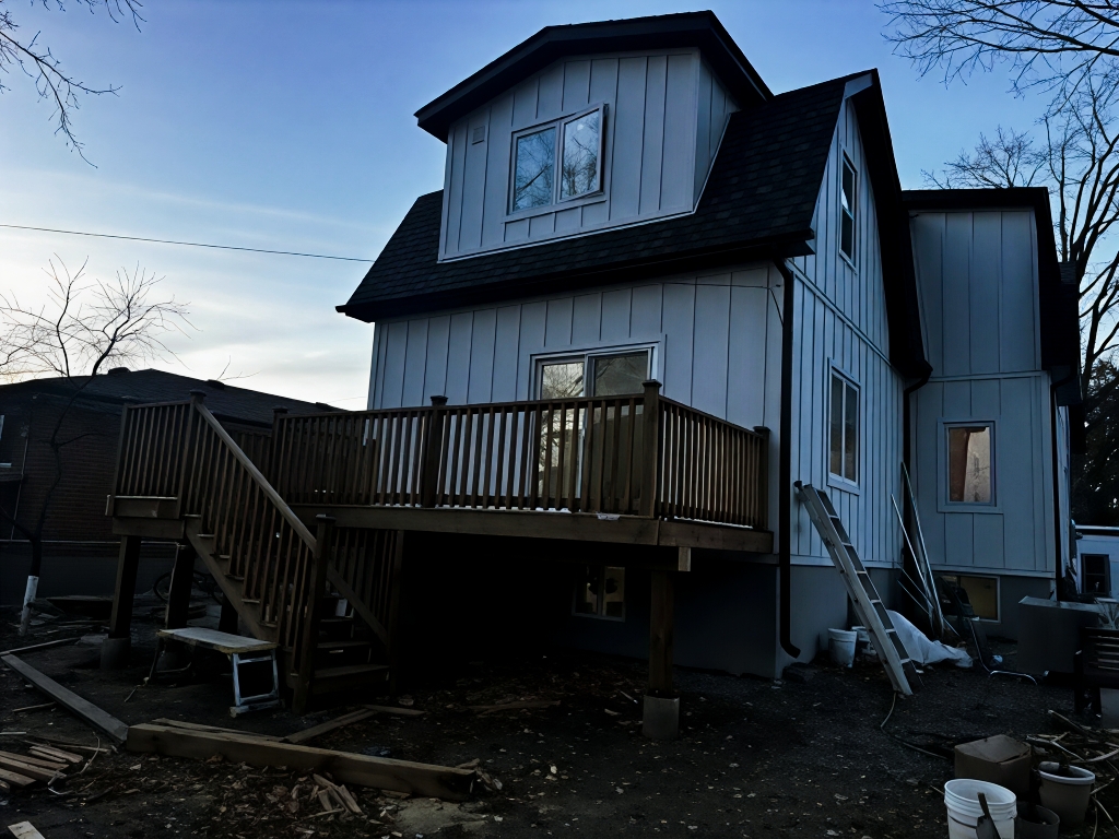 Residential house exterior with a second-floor wooden deck, stairs, railing, vertical siding, and construction materials visible below.