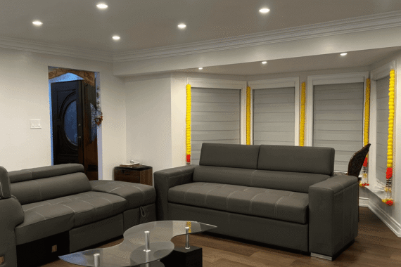 Modern living room with sectional sofas, hardwood flooring, glass coffee table, recessed ceiling lights, and window blinds.